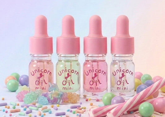 Four small bottles with pink droppers labeled 'Unicorn Oil mini' surrounded by colorful candies on a light background.