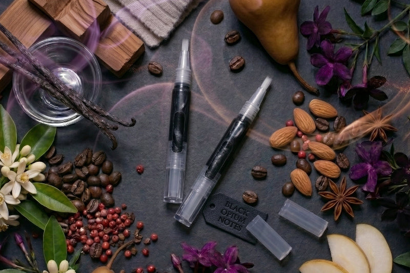 Midnight-inspired Unicorn Oil scent pens arranged with coffee beans, vanilla pods, pear, florals, almonds, pink peppercorns, and smoky purple accents for a Black Opium-inspired fragrance theme.