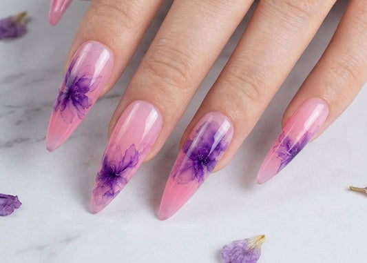 Hand with pink and purple floral nail design on a marble background with scattered flowers.